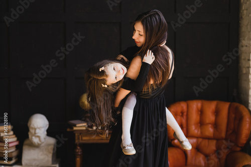 Mother and daughter hugging and playing together. Pretty little girl on beautiful woman's lap. Girls in black dresses playing in decorated room. Family weekend, beauty day, having fun, love concept.