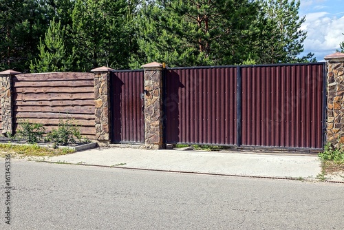 серо коричневые железные  ворота и деревянный забор перед асфальтированной дорогой 
