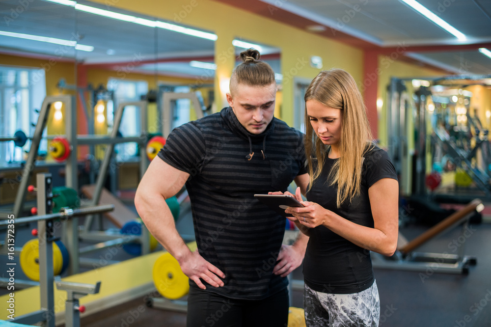 fitness, sport, exercising, technology and diet concept - smiling young woman and personal trainer with smartphone in gym