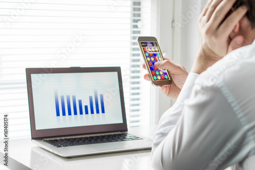 Lazy office worker playing mobile game with smartphone during work hours. Avoiding his job and leaning head against hand. Bored employee. Useless and unproductive man doing nothing and forget his job.