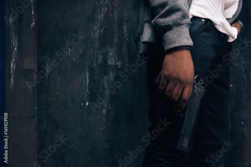 Unrecognizable armed black rapist closeup studio shoot. Gangster man with gun in hand on dark background. Outlaw, ghetto, murderer, robbery concept