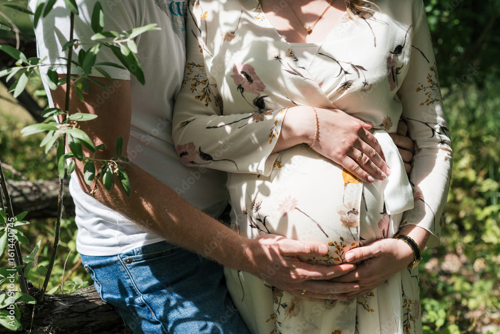 Close-up of human couple hands holding pregnant belly outdoors. Husband tenderly hugging pregnant wife in dress with floral pattern. Close up of loving pregnant couple