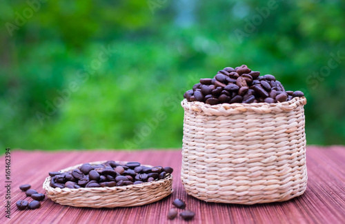 Good coffee beans are in a rattan basket. And some are in the lid on the bamboo mat. Close up and blurred green nature background.
