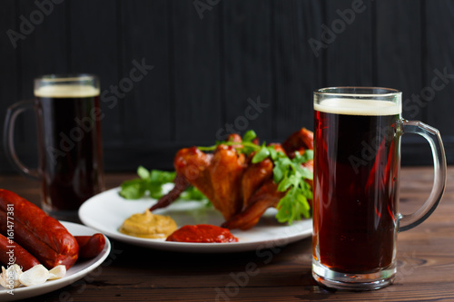 Two mugs of dark beer with crusty grilled chicken wings and various sausages.