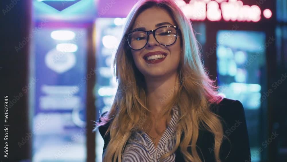 Close up view of a pretty blonde woman in formal suit, glasses looking right towards the camera and smiling happily at night. Bright city lights on the background. Cheerful mood, being happy. Close up view of a pretty blonde woman in formal suit, glasses looking right towards the camera and smiling happily at night. Bright city lights on the background. Cheerful mood, being happy.