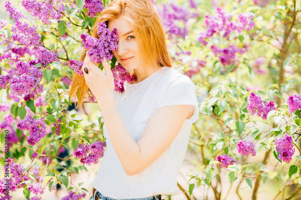 Fototapeta premium Portrait of young attractive cute redhair girl outdoor in park in blooming lilac bushes in summer sunny day. Resting among paradise flowers. Fragrance and fresh smell. Tenderness, innocence, airness.
