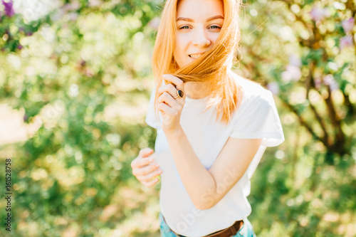 Young beautiful emotional tender redhead cutie girl getting fun and smiling. Playing with hair, looking at camera in park outdoor in summer sunny day on weekend with bright abstract background.