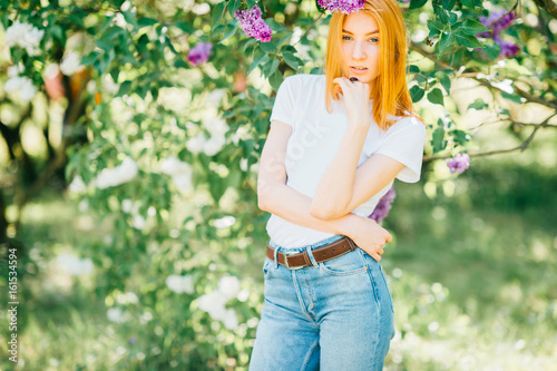Beautiful and attractive cute redhead girl in casual clothes posing for camera in exotic park outdoor with lilac bushes in summer sunny day. Blue jeans with belt. Youth fashion style. Selfaffirmation.
