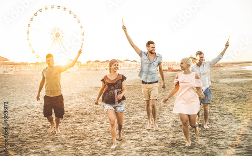 Group of happy friends having fun on the beach at sunset - Summer joy and friendship concept - Warm sunshine filtered color tone with focus on tall guy in the middle holding sparkle bengal fire candle