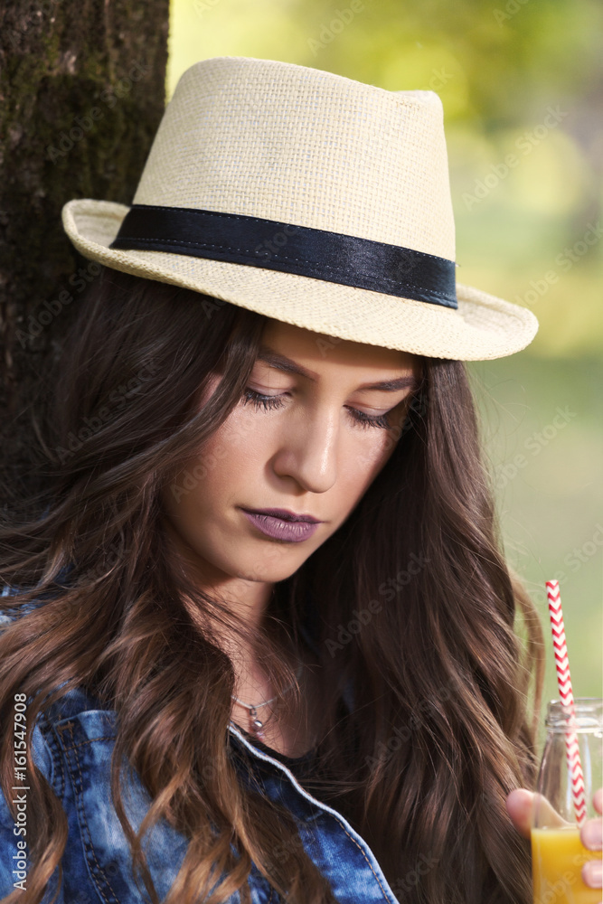 Beautiful young woman with a hat sitting in a park, holding a bottle with her favorite juice and relaxing