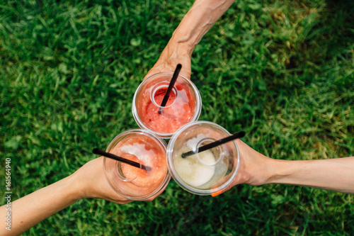 Three woman's hands holding plastic cup with smoothies over green grass background. Generation, sport, frienship and healthy life concept photo