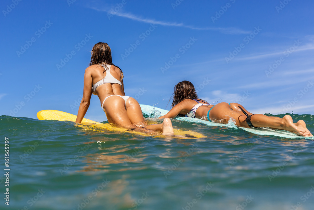 Two beautiful surfing girl with sexy fit butt on surf longboard surfboard board on sunrise or sunset in the ocean. Friends swim to lineup thru waves. Modern active sport lifestyle and summer vacation.