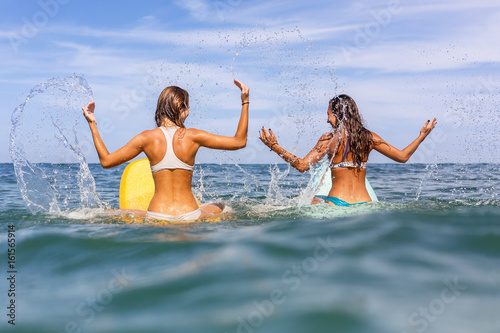 Two beautiful sporty surfing girl in sexy bikini sit on a big longboard surf surfboard board and make splashes on sunrise or sunset in the ocean. Modern active sport lifestyle and summer vacation.