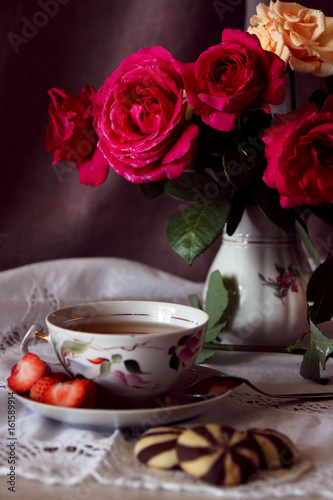 A cup of tea and biscuits against a vase with a bouquet of roses..
