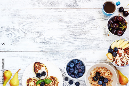 Healthy summer breakfast food frame. Top view of oatmeal, puffed rice cereal, pancakes with blueberry, cherry and pear, cup of black coffee on white wooden table with copy space. 