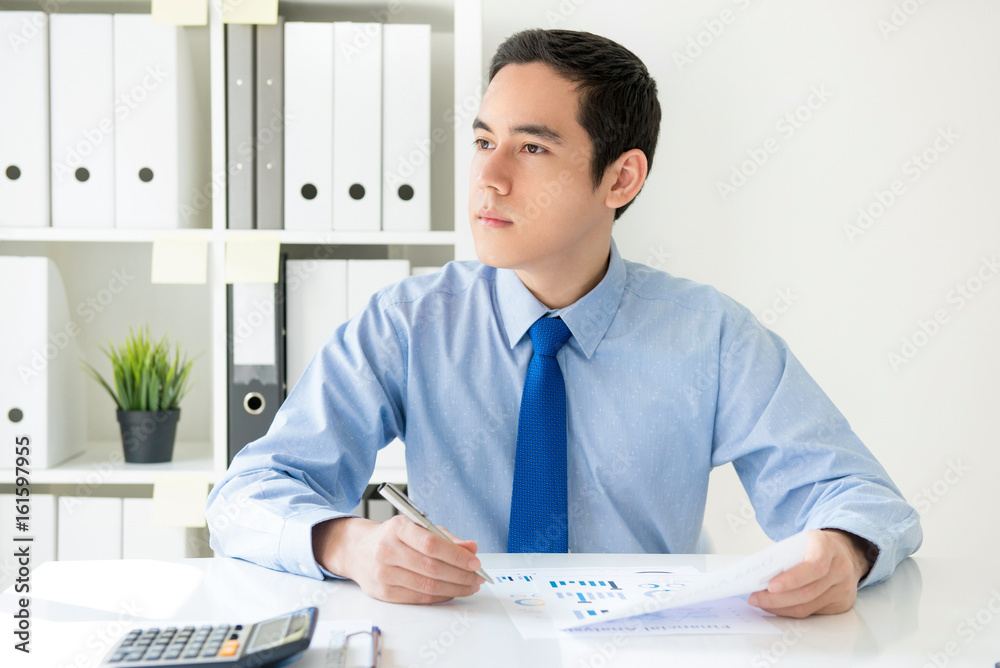 Young businessman looking out and thinking while sitting at working desk in the office Young businessman looking out and thinking while sitting at working desk in the office