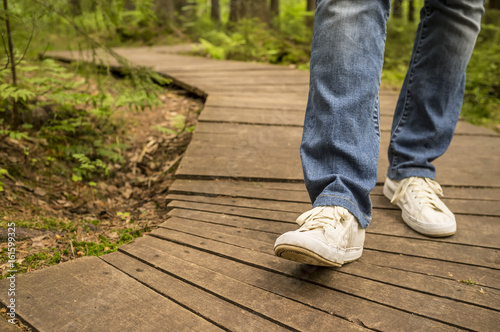 Girl in white sneakers and jeans walking along ecological wooden path in the summer forest.