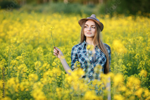 Attractive young blonde woman in blue plaid shirt straw hat enjoying her time on bright colorful blossoming yellow green meadow hard noon sun. © sergeyzapotylok