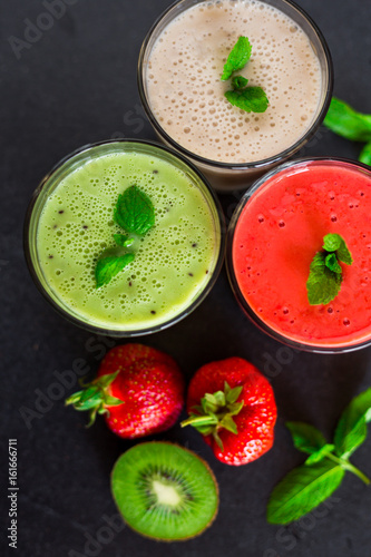 Close-up three glasses with smoothies, banana, kiwi, strawberries, on a black stone surface, decorated with mint, next to lie fresh berries and fruits, a tasty bright dessert, healthy food, restaurant