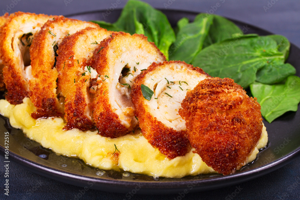 Chicken Kiev on mashed potato with spinach. Breaded chicken breast stuffed with herbs and butter on black plate Chicken Kiev on mashed potato with spinach. Breaded chicken breast stuffed with herbs and butter on black plate