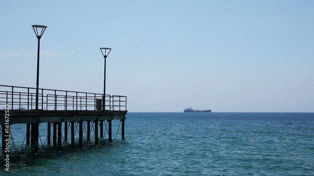 View of a pier with lamp posts, blue sea waves and a distant ship on the background View of a pier with lamp posts, blue sea waves and a distant ship on the background