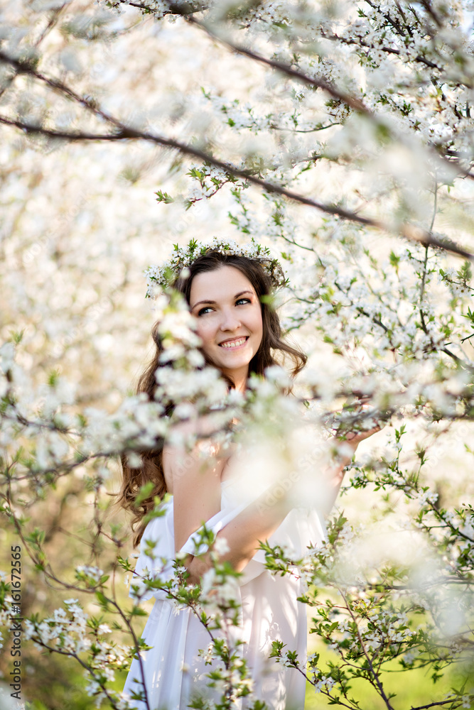 Beautiful young girl in white dress and wreath of flowers on head in blooming gardens Beautiful young girl in white dress and wreath of flowers on head in blooming gardens