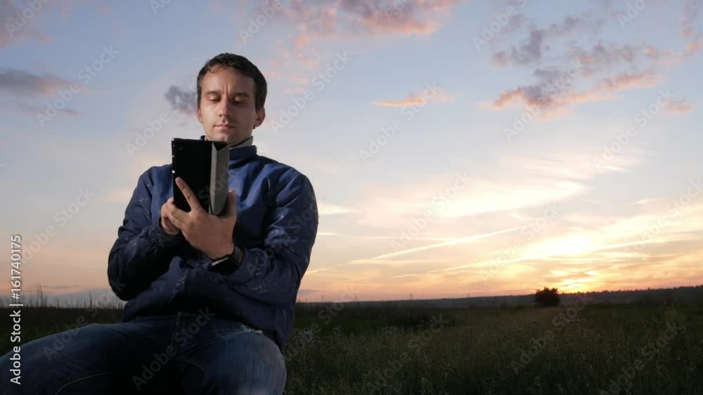 A young man sits at sunset and checks the messages to check the messages on the tablet. Beautiful sky behind. A young man sits at sunset and checks the messages to check the messages on the tablet. Beautiful sky behind.