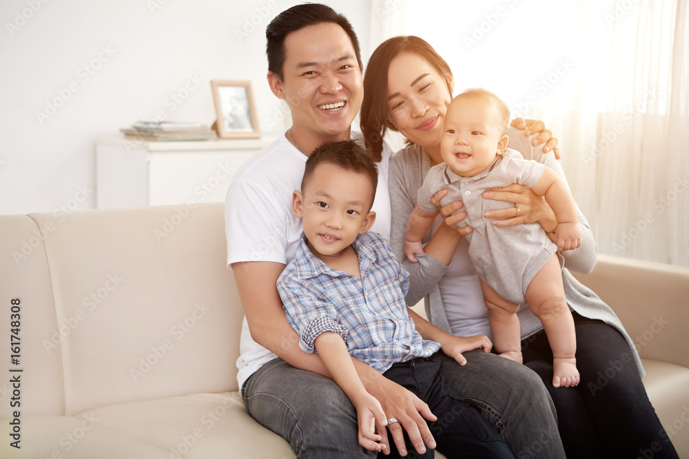 Portrait of happy Asian family with two children posing on sofa at home looking at camera in sunlight Portrait of happy Asian family with two children posing on sofa at home looking at camera in sunlight