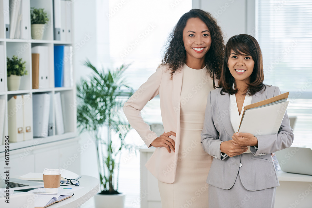 Portrait of two attractive middle-aged businesswomen looking at camera with wide smiles while standing in modern open plan office Portrait of two attractive middle-aged businesswomen looking at camera with wide smiles while standing in modern open plan office