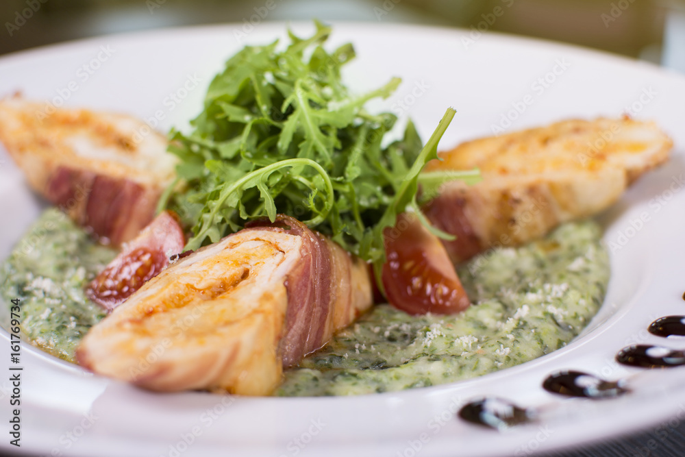 Roasted chicken breast wrapped in bacon slices with greens, tomatoes, and sauce. Beautifully served meal at the restaurant.