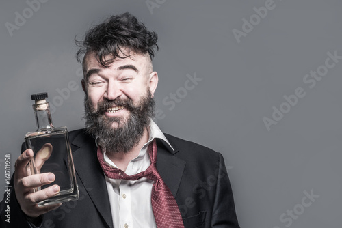 Tableau sur toile Disheveled smiling bearded man in suit holding in his hand a bottle of alcohol o
