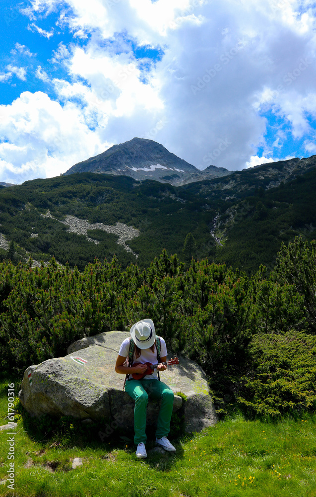 Tourist is playing on the Ukulele guitar at the mountain nature green landscape. Photo depicts romantic view of the young man playing on musical instrument in the mountains.