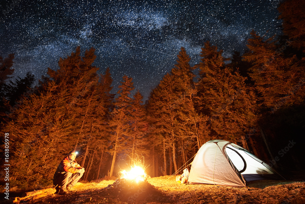 Male tourist have a rest in his camp near the forest at night. Man sitting near campfire and tent under beautiful night sky full of stars and milky way, and enjoying night scene - obrazy, fototapety, plakaty 