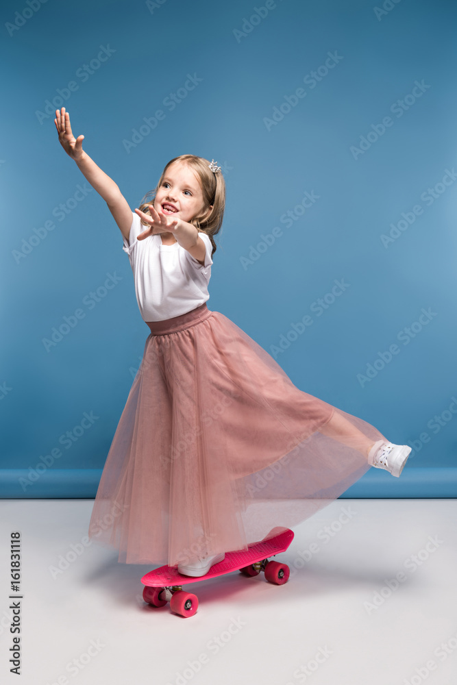 Naklejka premium Cute smiling girl in pink skirt standing on skateboard and having fun