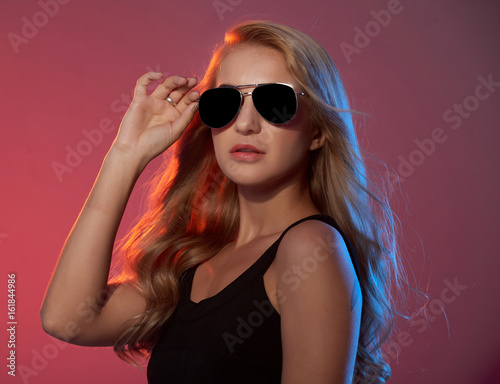Young blonde-haired fashion model wearing black top and sunglasses posing against red background