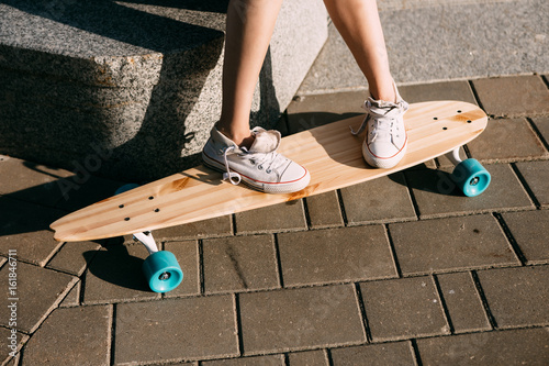 Close up of lady leg in white sneakers resting after extreme funny ride her wooden longboard skateboard. Modern urban hipster girl have fun. Good sunny summer day for skateboarding and have fun.
