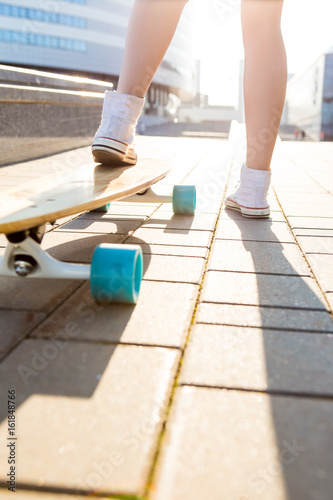 Close up of sporty woman in white sneakers ready to extreme funny ride her wooden skate longboard skateboard in sun light. Modern urban hipster girl have fun. Good sunny summer day for skateboarding.