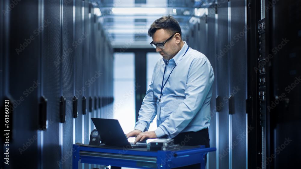 In Data Center IT Engineer Works on Crash Cart Laptop. We See Rows of Server Racks and One Cabinet is Open. In Data Center IT Engineer Works on Crash Cart Laptop. We See Rows of Server Racks and One Cabinet is Open.
