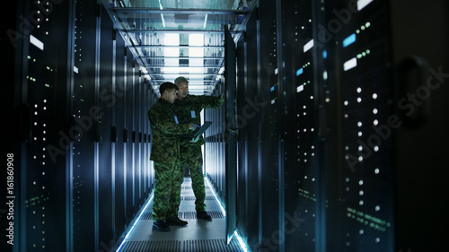 In Data Center Two Military Men Work with Open Server Rack Cabinet. One Holds Military Edition Laptop. photo