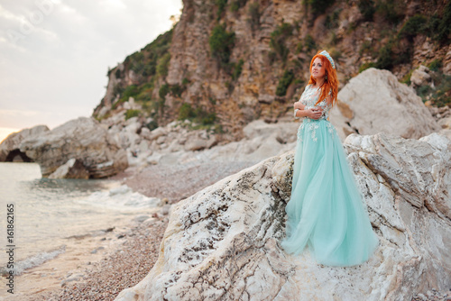 Young beautiful red-haired woman in a luxurious dress stands on a rocky shore of the Adriatic Sea among large stones