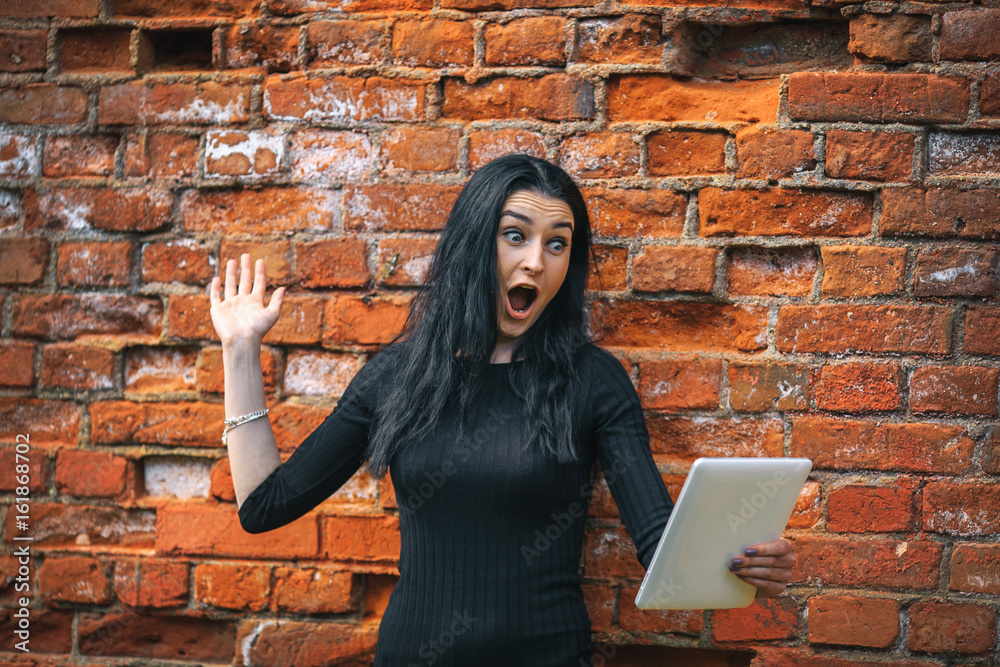 Gadget woman. Portrait of surprised young pretty white girl with long black hair and in black clothes surfing on the internet in white tablet near vintage red brick wall. Gadget woman. Portrait of surprised young pretty white girl with long black hair and in black clothes surfing on the internet in white tablet near vintage red brick wall.