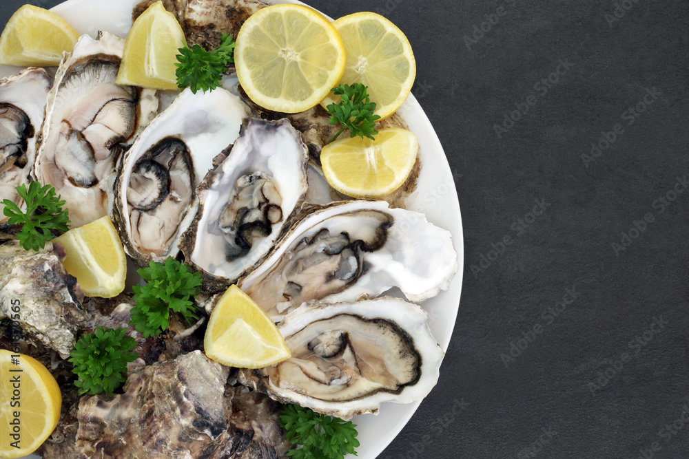 Oyster aphrodisiac food on a porcelain plate with lemon fruit and parsley herb on slate background.