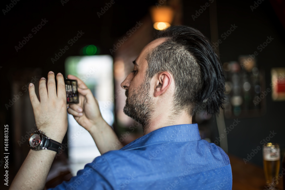 Fototapeta premium Young bearded man taking photos with his mobile phone in the bar