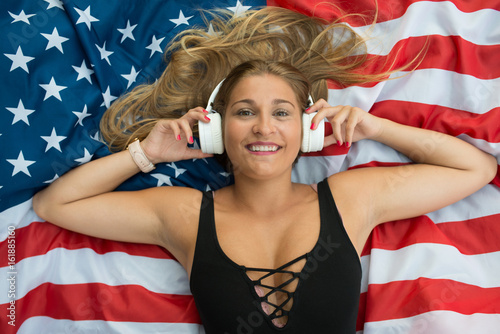 American blonde woman resting on the grass with the American flag listening to her playlist with her wireless headphones