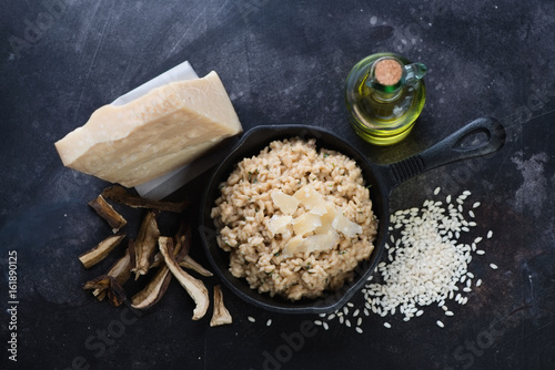 Risotto with ceps in a frying pan and its cooking ingredients over dark scratched metal background, above view