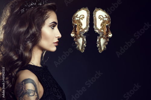 Close up portrait of charming lady with dark wavy silky hair and perfect make-up wearing jewel crown and looking in the face of Venetian mask hanging in the air. Backlight. Copy-space. Studio shot photo