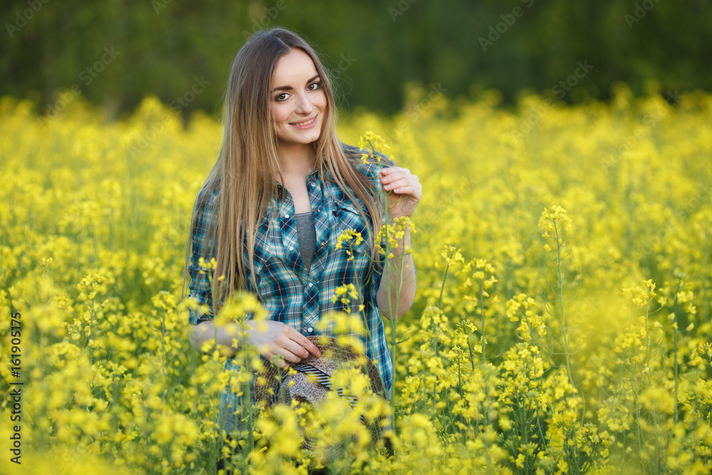 Attractive young blonde woman in blue plaid shirt straw hat enjoying her time on bright colorful blossoming yellow green meadow hard noon sun. Attractive young blonde woman in blue plaid shirt straw hat enjoying her time on bright colorful blossoming yellow green meadow hard noon sun.