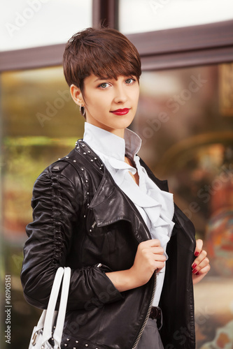 Happy young fashion woman in leather jacket with handbag next to mall window