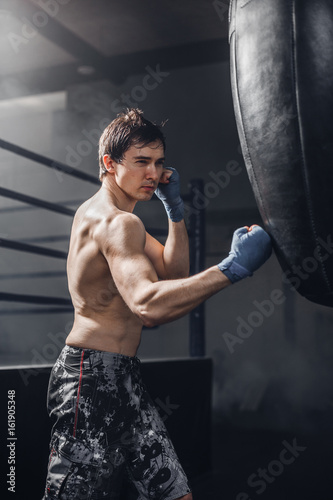 Muscular fighter prepare for practicing some kicks with punching bag. Muscular strong man on background boxing gym. © belyjmishka