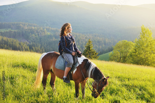 Young woman sits on a horse in the mountains at sunset in summer. Active holiday in the mountains on vacation.
